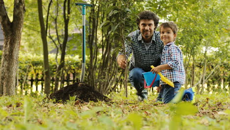 Portrait-of-a-little-boy-and-his-dad-planting-a-tree.-They-look-into-the-camera-and-smile.-Then-the-boy-plays-and-dad---touches-his-son's-hair.-Blurred-background